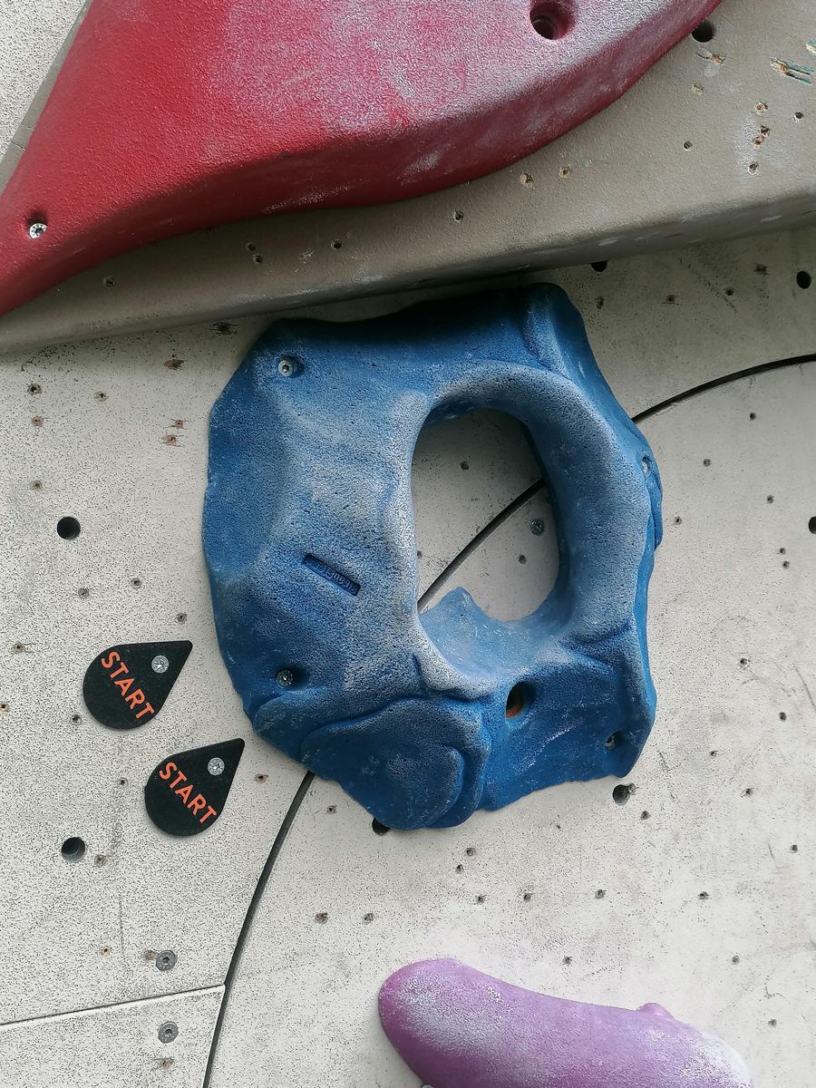 Zwei tropfenförmige Startschilder neben einem blauen Griff an der Boulderwand